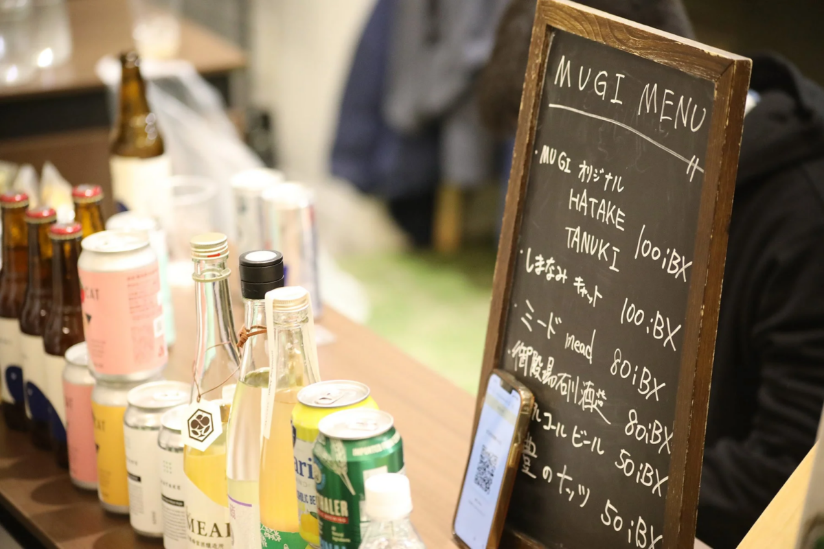 カウンターに並べられた様々なボトルや缶の飲み物と、手書きのMUGIメニューが書かれた黒板が写っています。メニューにはオリジナルドリンクやビール、ミードなどが表示されており、QRコード付きのスマートフォンも置かれています。