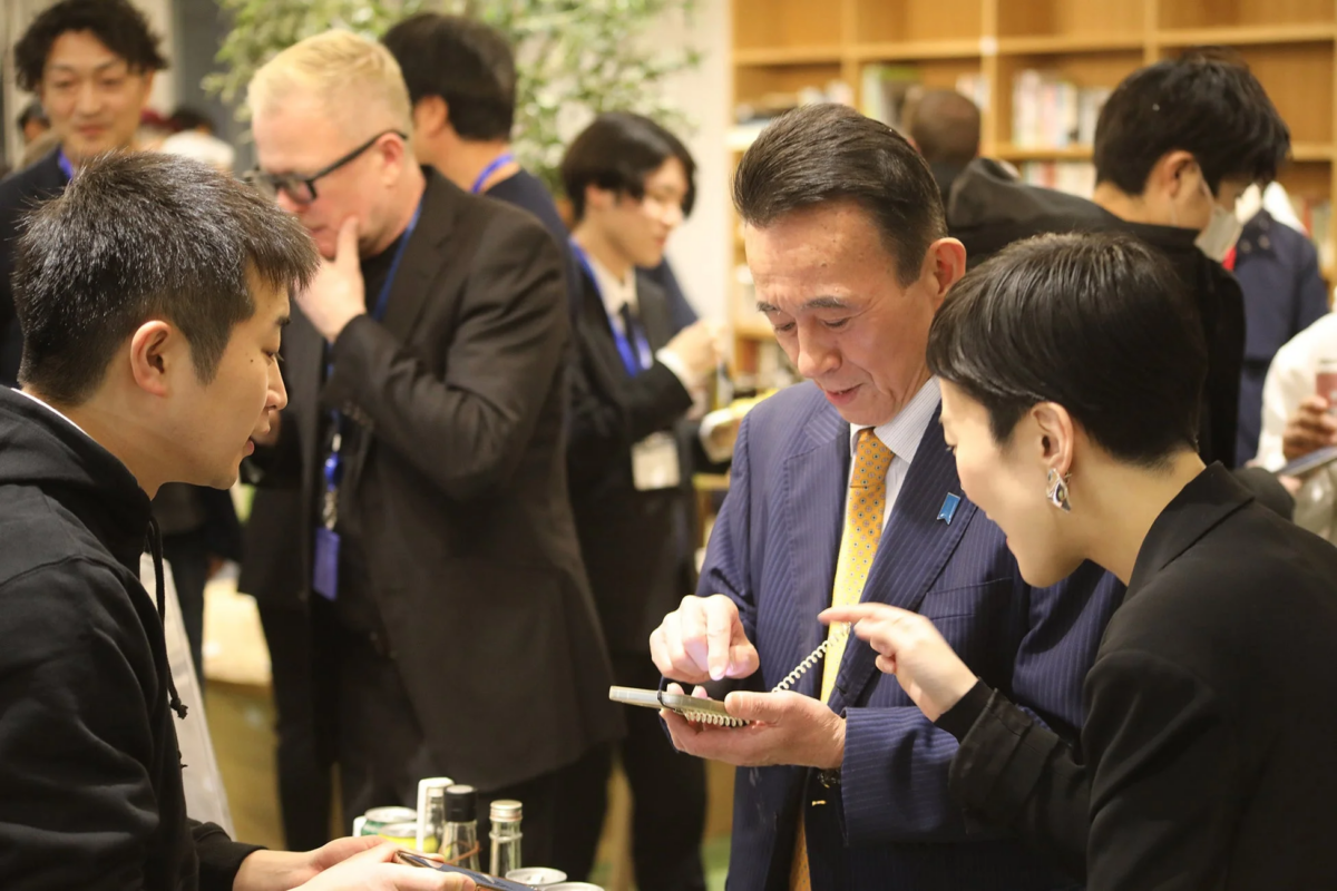 屋内のイベントで、スーツ姿の男性がスマートフォンを操作し、隣の女性が画面を指差して説明している様子。