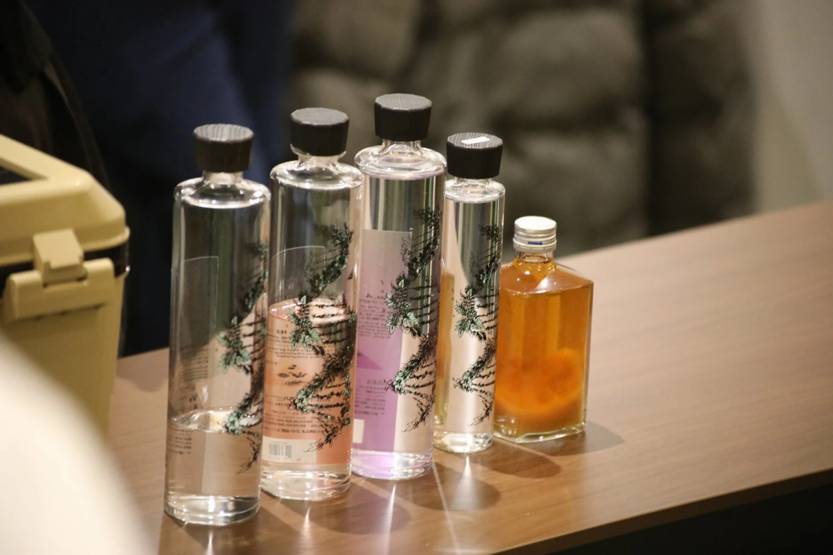 木目調のテーブルに様々な形状のガラスボトルが5本並べられています。透明なボトルには植物のデザインが施され、液体が満たされています。一番右のボトルは琥珀色の液体が入っており、化粧品やアロマ製品のような展示風景です。