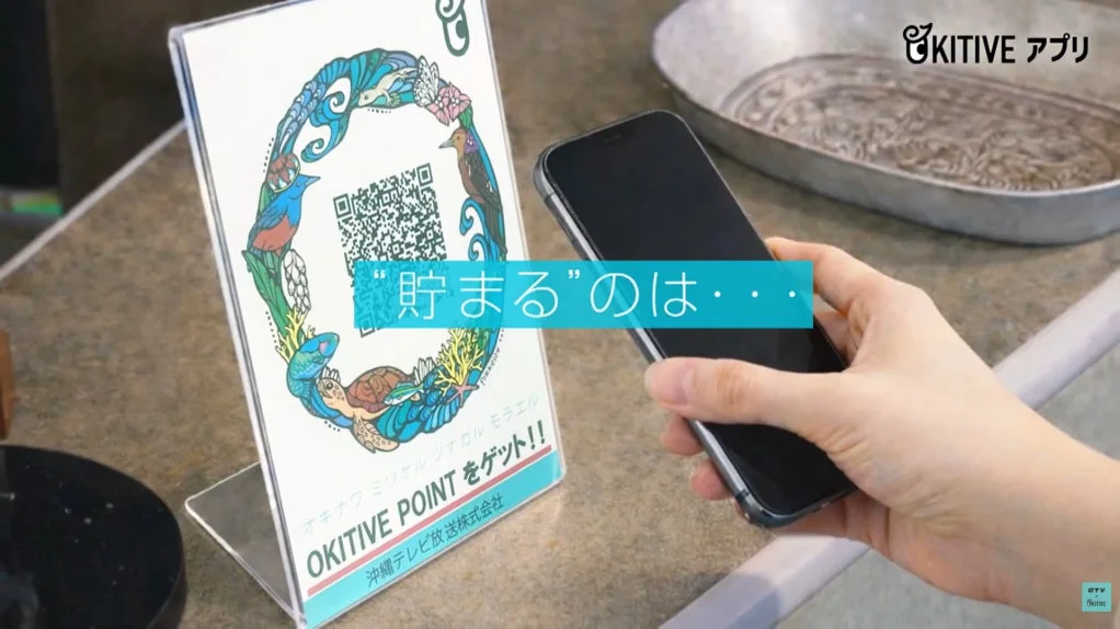 スマートフォンでOKITIVEアプリのQRコードをスキャンし、ポイントを獲得するシーンです。沖縄の自然をモチーフにしたデザインが特徴的なQRコードスタンドが映っています。