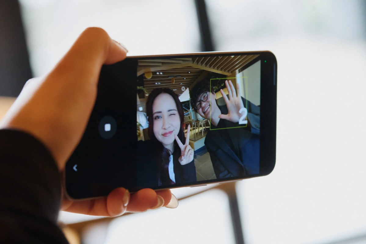 スマートフォンで自撮りをする男女の様子。女性はウィンクとピースサイン、男性は笑顔で手を広げ、画面には数字の「2」が表示されており、撮影の瞬間の楽しげな雰囲気が伝わります。