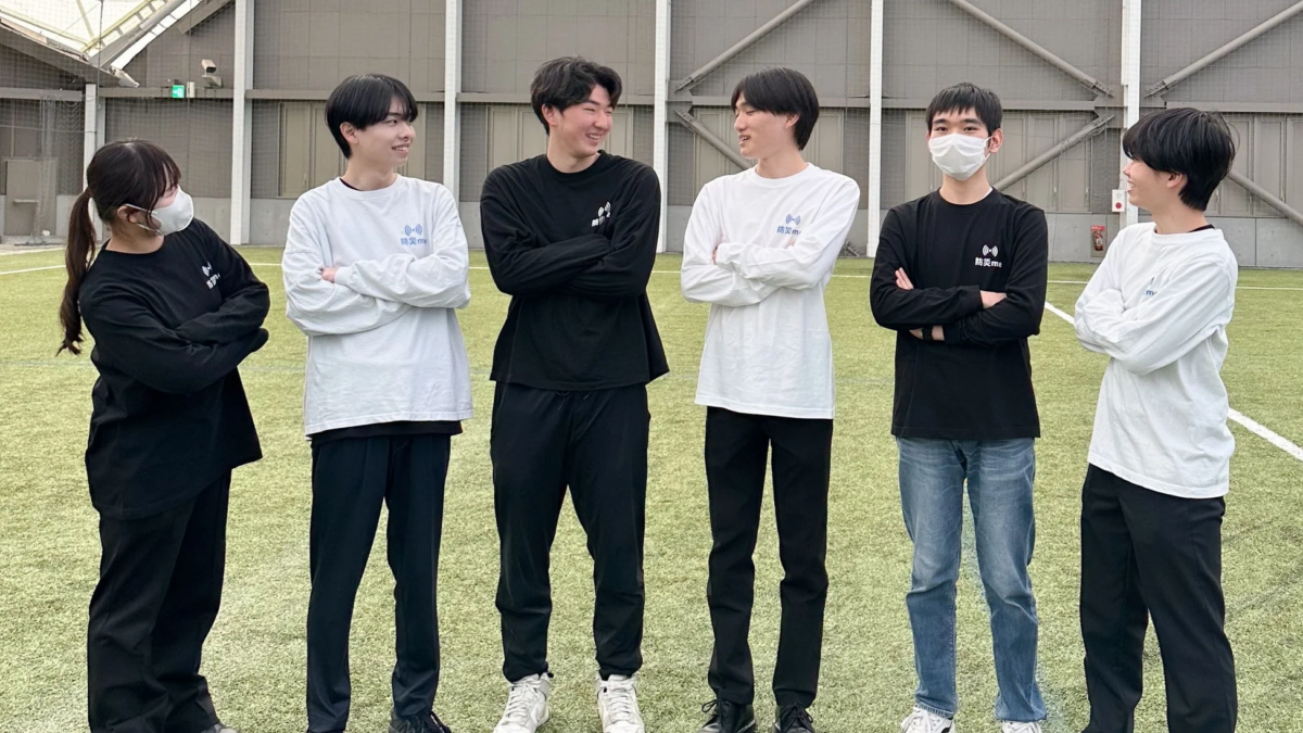 「防災me」のロゴ入りシャツを着た6人の若者が、屋内の人工芝の上で笑顔でポーズをとっている集合写真です。チームやプロジェクトメンバーの記念撮影のようです。