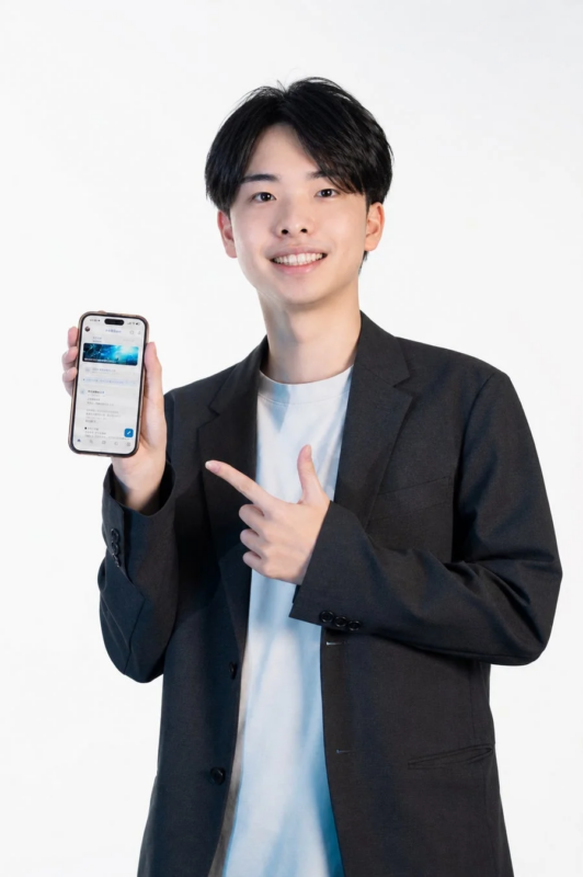 白い背景の前で、笑顔の若い男性がスマートフォンを持ち、その画面を指差しています。彼は黒いブレザーを着用しており、スマホの画面にはアプリの内容が表示されています。