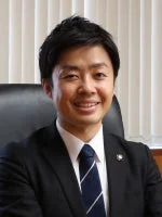 スーツを着用し、ストライプのネクタイを締めたアジア系の男性がカメラに向かって笑顔で写っている