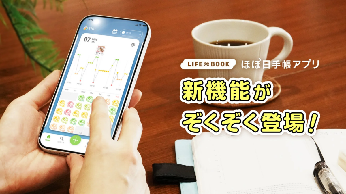 スマートフォンで「LIFE@BOOK ほぼ日手帳アプリ」を使用している様子。グラフや日付、気分アイコンが表示され、新機能が続々登場することをアピールするテキストが大きく表示されています。手元にはコーヒーカップとノートも置かれ、落ち着いた環境での利用が示唆されます。
